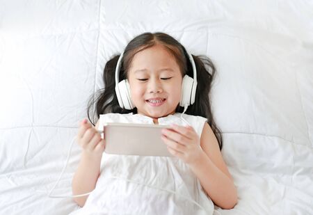 Happy little Asian girl using headphones listen music by smartphone smiling while lying on bed at home.の写真素材