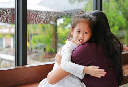 Asian mother and her daughter hugging with love near a glass window while raining day. Cute little girl looking at camera with smiling.の写真素材