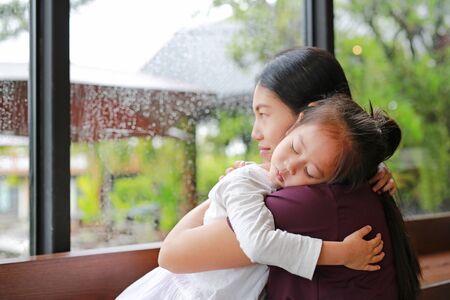 Asian mother and her daughter embracing with love near a glass window while raining day.