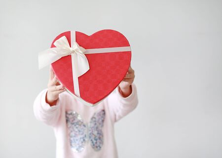 Adorable little Asian child girl showing red heart gift box on white background. Kid giving red heart gift box for you. Concept of love.の写真素材