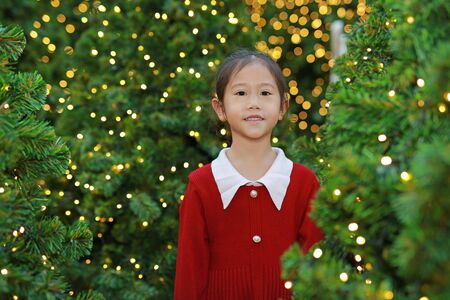 Happy Asian little girl in red dress against Christmas tree in a background.の写真素材