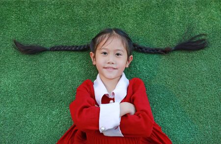 Beautiful Asian little girl in red dress cross one's arm and lying on green grass background in the winter season and happy new year.の写真素材