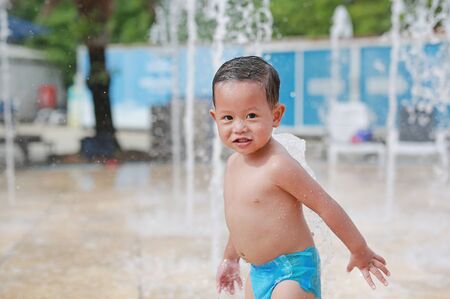 Happy little Asian baby boy having fun on water stream of a sprinkler. Kid playing in playground fountain in aqua park.の写真素材
