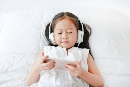 Portrait little Asian girl using headphones listen music by smartphone while lying on bed at home.の写真素材