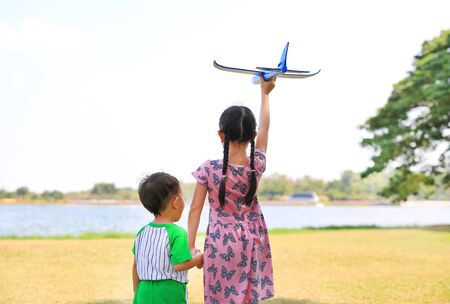 Asian little child girl hold hands baby boy and raise up a blue toy airplane flying on air in the nature garden. Sister and her brother standing near lake in the park. Rear view.の写真素材