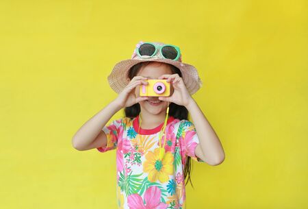 Asian little child girl wearing a floral pattern summer dress and hat with sunglasses taking photo by toy camera at you isolated on yellow background. Holiday and summer fashion concept.の写真素材