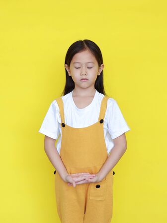 Peaceful asian little girl with closed eyes standing isolated on yellow background. Kid in meditation expressionの写真素材