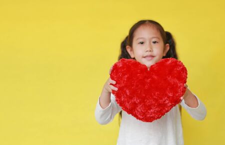 Smiling asian child girl holding a fluffy red heart for you on yellow background. Selective focus at red heart.の写真素材