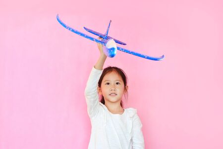 Cheerful asian little child girl raise up a blue toy airplane flying on air isolated on pink background.の写真素材