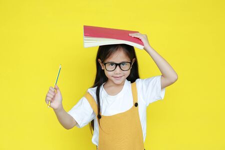 Happy little girl back to school concept. Kid with book on head and holding pencil with smiling...