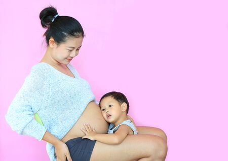 Asian baby boy listening baby in tummy of pregnant mother isolated on pink background. Pregnancy and new life conceptの写真素材
