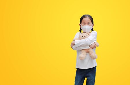 Coronavirus covid-19 and pollution protection concept. Asian little child girl hugging teddy bear doll with wearing mask to against corona virus and air pollution pm2.5 isolated on yellow backgroundの写真素材