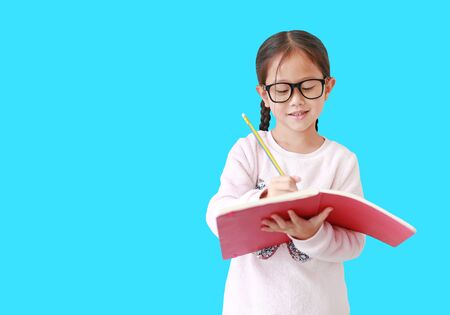 Asian girl wearing eyeglasses holding and writes in notebook with pencil isolated on light blue backgroundの写真素材
