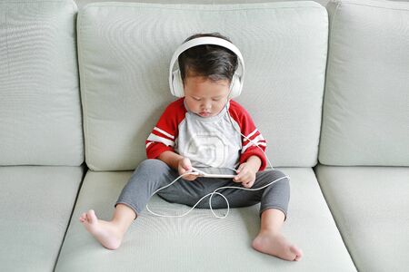 Cute little Asian baby boy in headphones is using a smartphone lying on the sofa at home. Child listening to music on earphones.の写真素材
