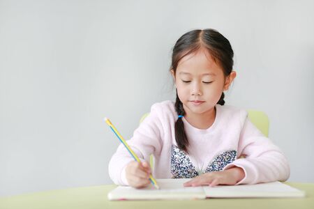 Portraits of little Asian child girl write in a book or notebook with pencil sitting on kid chair and table against white background.の写真素材