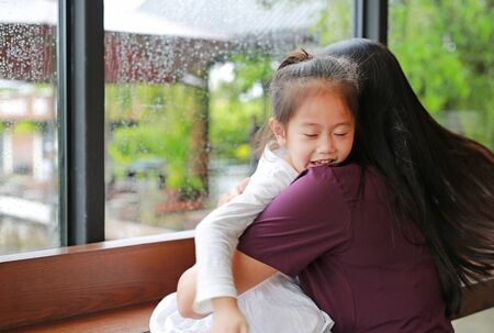 Beautiful Asian mother and her daughter embracing with love near a window while raining day.