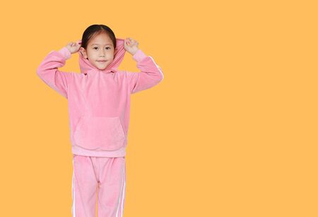 Portrait of little Asian child girl in pink tracksuit or sport cloth with putting hood on head isolated on orange background with copy space. Portrait half-length of kid girl.の写真素材