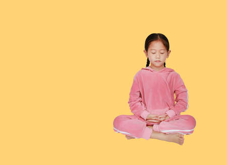 Cute little asian child girl in pink tracksuit with eyes closed practicing mindfulness meditation sitting isolated over yellow background with copy space. Peaceful concept.の写真素材