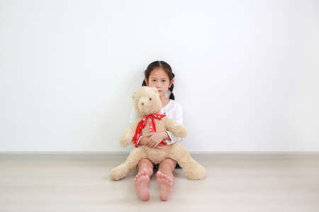 Peaceful asian little kid girl hugging a teddy bear doll sitting against white wall in the room alone.の写真素材