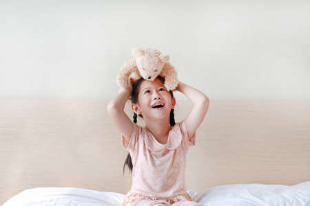Happy Asian little girl showing teddy bears while sitting on the bed at home.の写真素材
