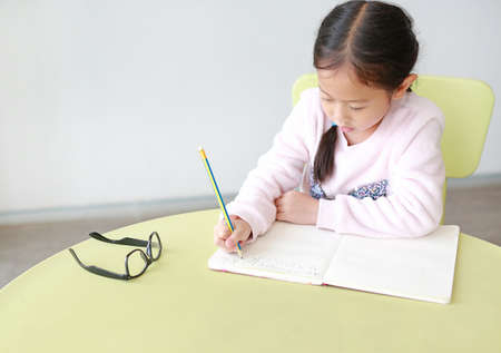 Portraits of little Asian girl writes in a book or notebook with pencil sitting on kid chair and table in classroom.の写真素材
