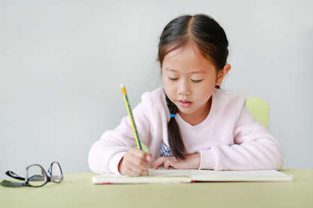 Portraits of little Asian child girl write in a book or notebook with pencil sitting on kid chair and table against white background.の写真素材