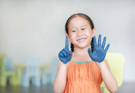 Happy little Asian girl with her blue hands painted showing one and five fingers in kids room.の写真素材