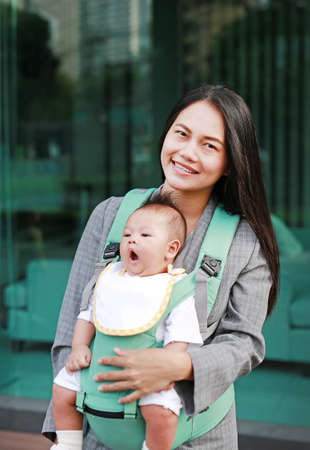 Young working mother carrying her infant by ergonomic baby carrier.