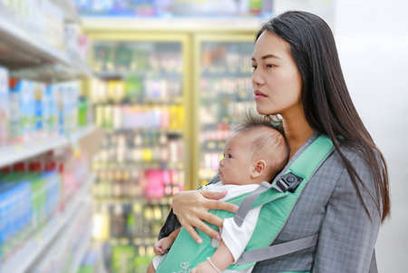 Young business woman in Convenience Store while carrying her baby boy by ergonomic baby carrier.