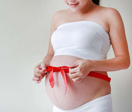 Portrait of pregnant woman tummy with tying red ribbon gift.の写真素材