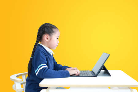 Asian little kid girl in school uniform using laptop on table isolated on yellow background, Studio shotの写真素材
