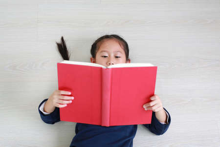 Close up asian little child girl in casual school uniform reading book and lying on wooden laminate flooring. Education conceptの写真素材