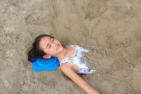 Portrait of happy asian little child girl buried in the sand at the beach. Close up kid playing with sand.の写真素材