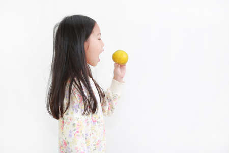 Portrait little asian kid girl holding not peeled orange fruit and gesture eating over white backgroundの写真素材