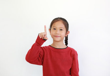 Smiling young asian little girl in red sweater pointing index finger up while looking at camera isolated on white background. Kid and advertising conceptの写真素材