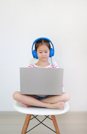 Asian little school girl sitting on chair using headphone study online learning class by laptop computer during new normal Covid-19 coronavirus at homeの写真素材