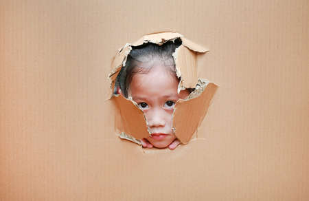 Adorable little Asian child girl looking through hole on cardboard.