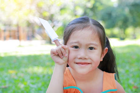 Happy little Asian child girl fold a piece of white paper to rocket and showing on her forefinger in the summer garden. Education and imagination concept.の写真素材