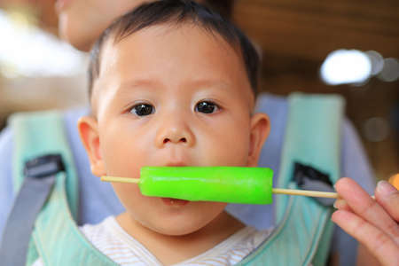 Adorable Asian baby boy sucking ice-cream by people hand.の写真素材