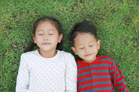 Top view Asian little child boy and girl lying on green grass outdoors in summer park. Portrait of sister and brother closed eyes resting in garden happy smiling togetherの写真素材