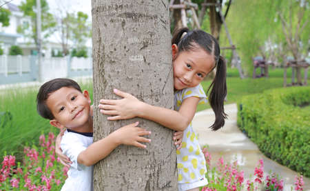 Portrait of cute little boy and girl child hugging with arms around tree trunk in the garden. Children enjoying nature life love, smiling in nature outdoors.の写真素材