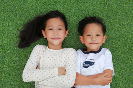 Asian little boy and girl child lying on green grass background together with looking camera. Above...