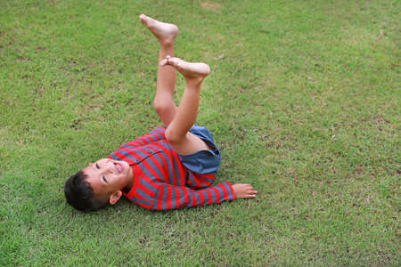 Adorable Asian little boy lying on grass with lift up his legs at the garden. Kid lies on green...