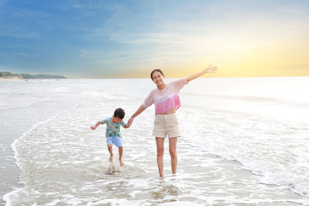 Cheerful Asian mother and little boy enjoy playing on tropical sand beach at sunrise. Happy family mom and son having fun in summer holiday.の写真素材