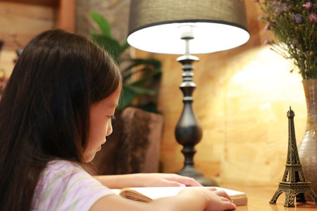 Close-up little Asian girl child in casual clothes reading a book and smiling on the desk in the room.の写真素材