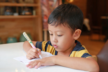 Intended Asian little boy drawing in a book with a pen on table in classroom.の写真素材