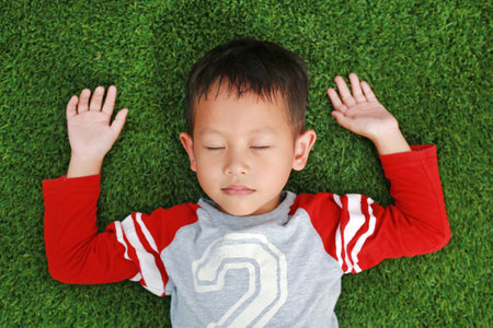 Peaceful Asian little boy close eyes and lying down on green lawn. Kid lies on grass. Top View.の写真素材