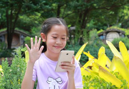 Asian girl kid speaking on video conference call on smartphone, waving greeting hand hello, talking to family online while in park outdoor. Distant communication concept. Child waving palm saying hi.の写真素材