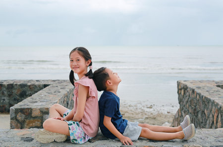Adorable Asian young girl child and little boy kid sitting together at seacoast. Sister and brother...
