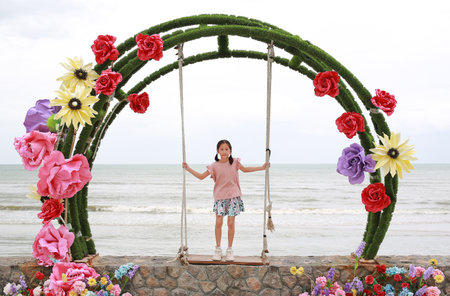 Asian young girl child standing on beautiful swing against cement wall at seaside. Full length.